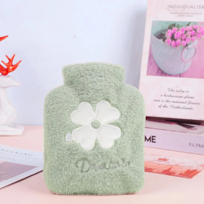 A light green plush-covered hot water bottle with a white flower design resting on a pink surface.