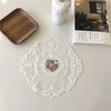 An elegant oval white lace embroidered placemat is displayed on a white table next to a wooden board and a white container.