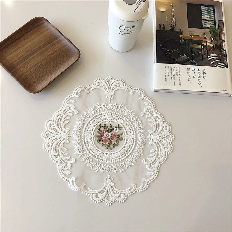 An elegant oval white lace embroidered placemat is displayed on a white table next to a wooden board and a white container.