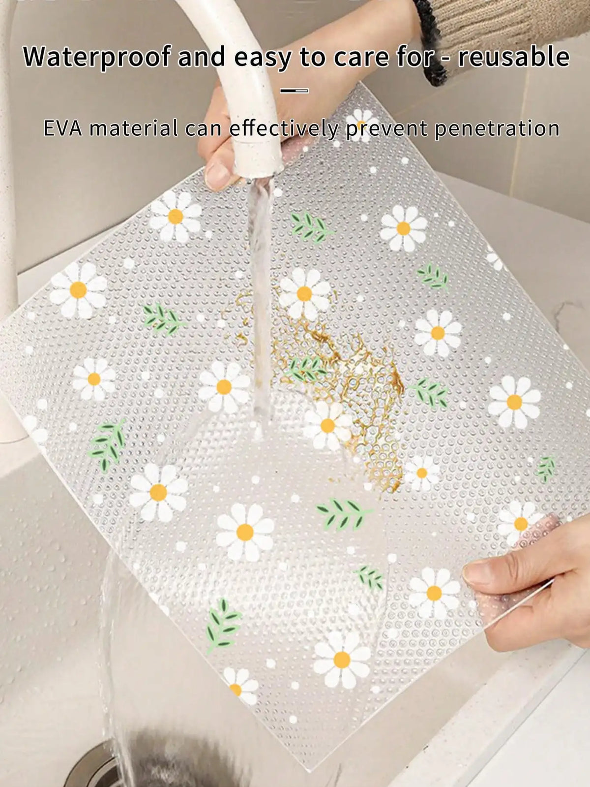 A clear, reusable EVA material mat with white daisy flowers and green leaves is being rinsed under water.
