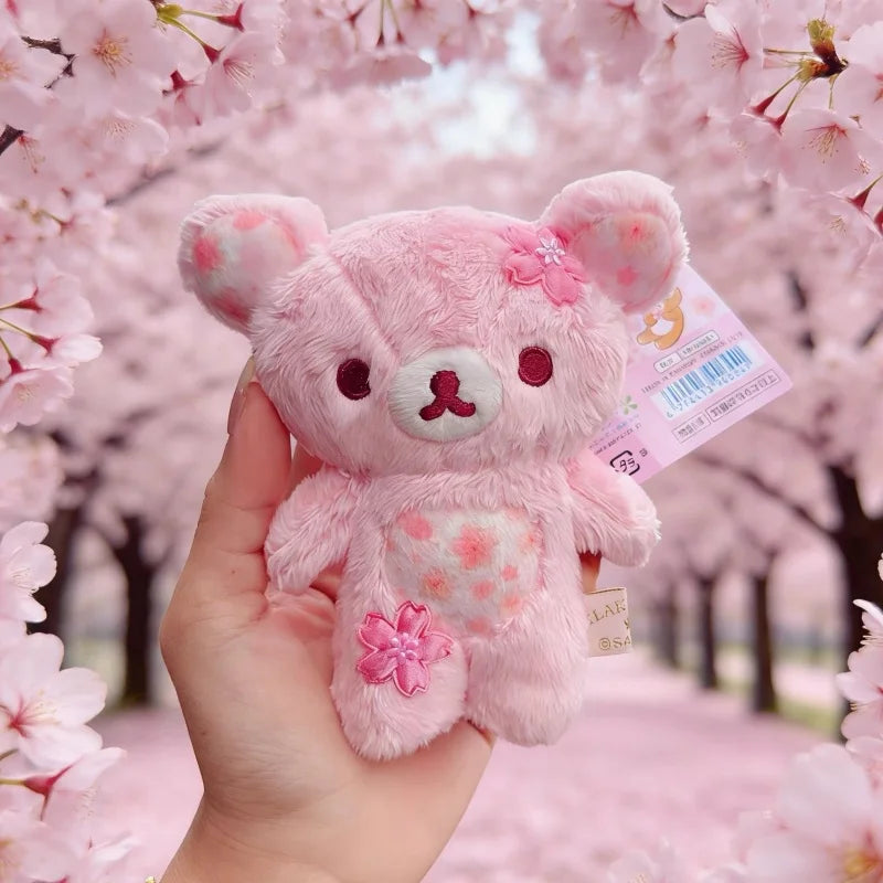 A pink cherry blossom plush bear keychain is held against a background of pink blossoms.