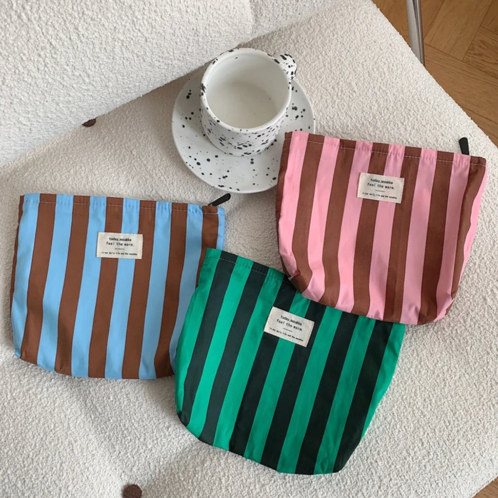 Three striped canvas cosmetic bags in blue, pink, and green are displayed on a white surface with a ceramic cup.