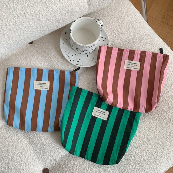 Three striped canvas cosmetic bags in blue, pink, and green are displayed on a white surface with a ceramic cup.