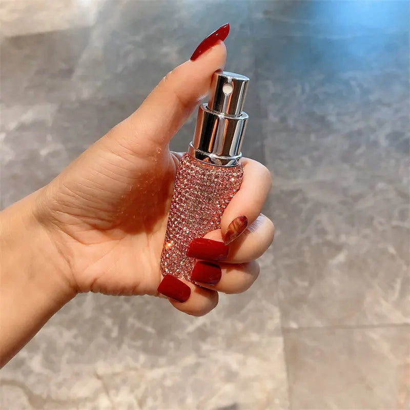 A hand holds a glass perfume atomizer filled with pink glitter and a silver spray top against a blurred background.