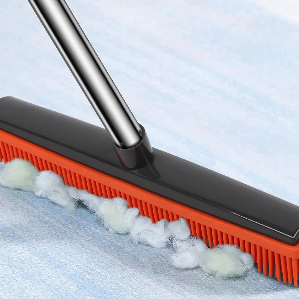A black rubber broom and carpet rake with an orange bristles is shown against a light blue background.