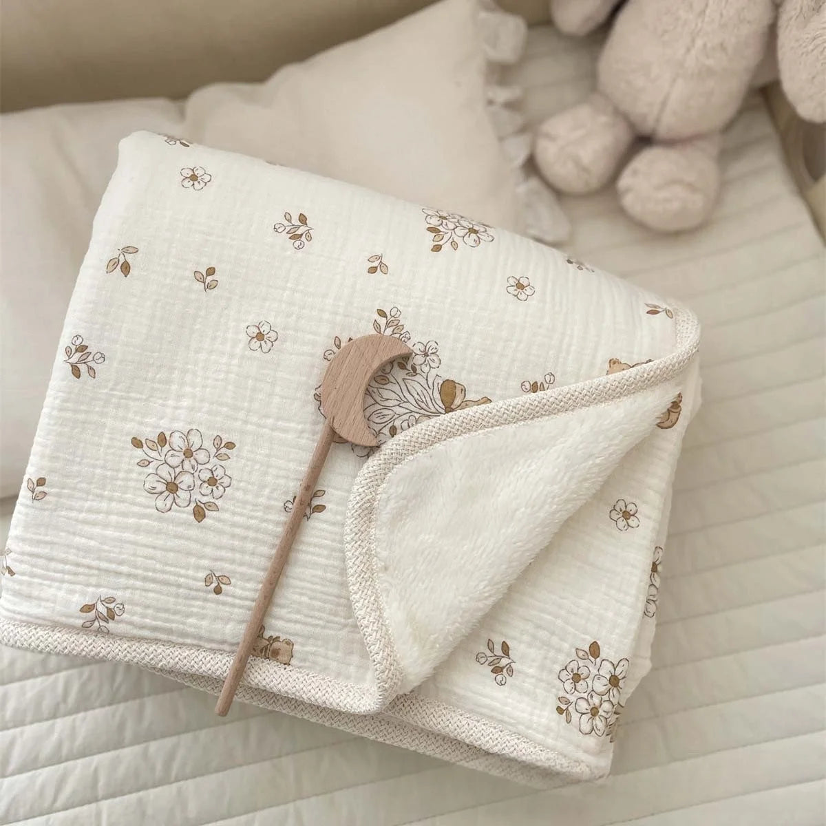 A white floral muslin baby blanket with a wooden moon shape is folded on a white textured surface.