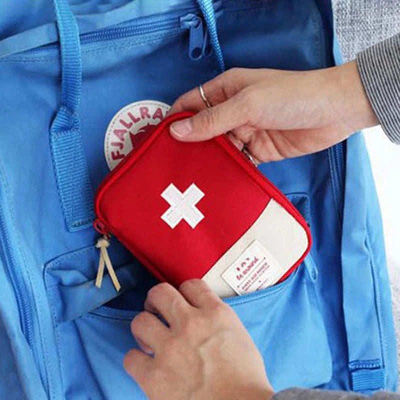 A hand is placing a red compact travel medicine organizer bag with a white cross on a blue backpack.