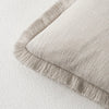 A beige linen throw pillow cover with a fringed edge resting on a white surface.