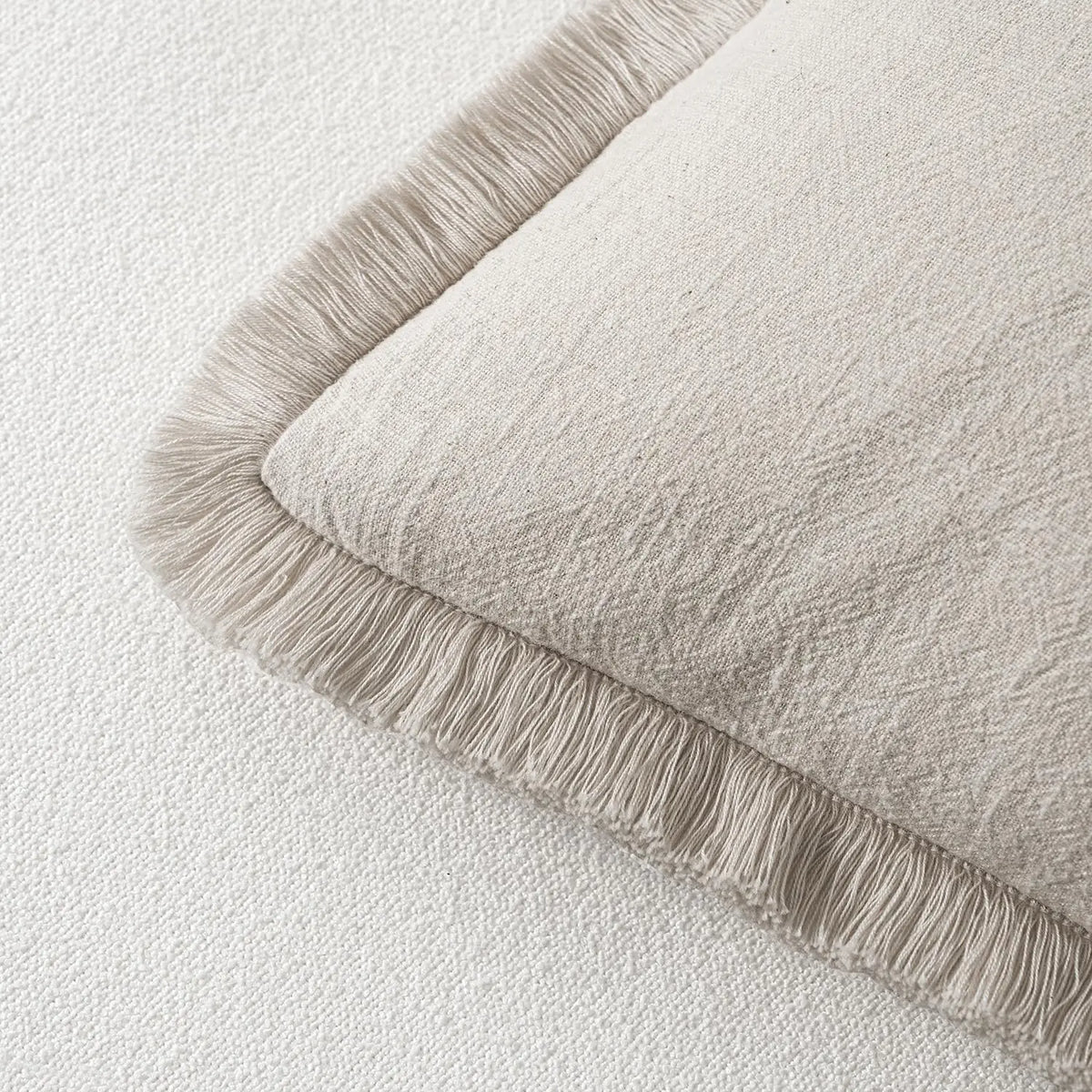 A beige linen throw pillow cover with a fringed edge resting on a white surface.
