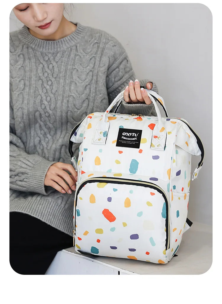 A woman holds a white and colorful patterned waterproof diaper bag backpack with black straps.