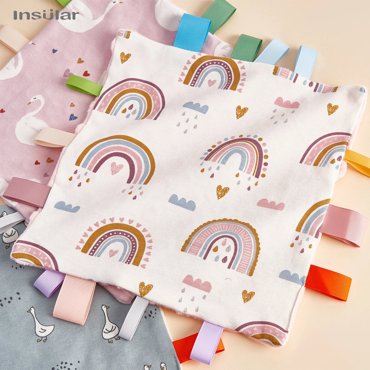 A white baby sensory tag comfort blanket with colorful rainbow patterns and clouds is shown on a light background.