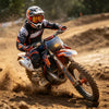A rider wearing orange and black gear is performing off-road motorcycle stunts on a dirt track.
