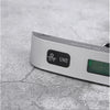 A silver portable digital luggage scale with a black unit button and an LCD display resting on a gray surface.