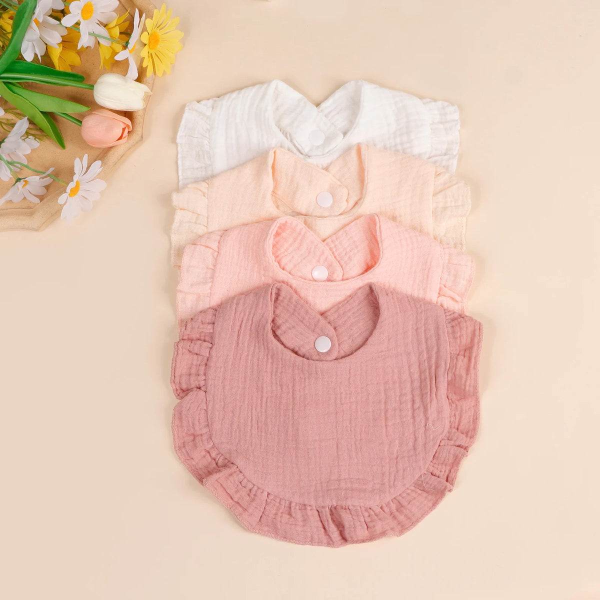 A set of three soft cotton ruffle bibs in shades of cream, peach, and dusty rose laid out on a light background.