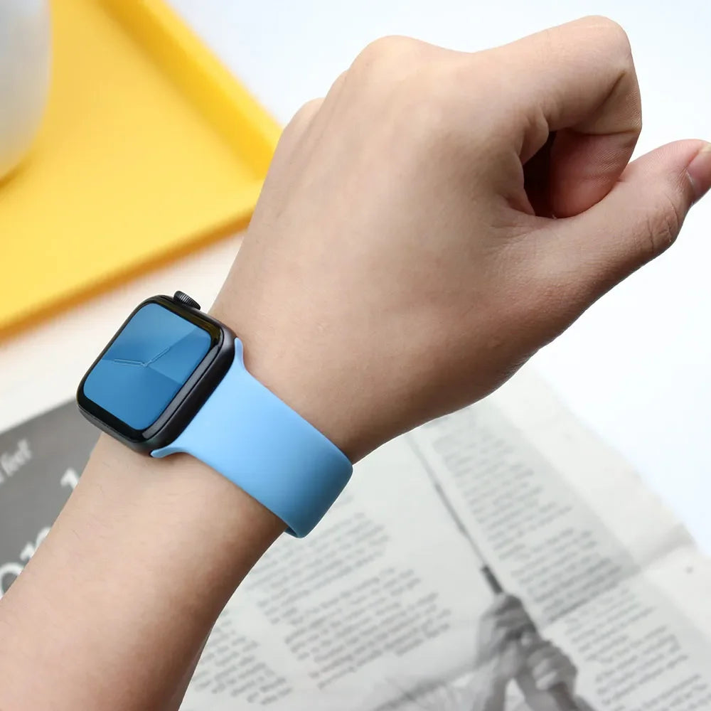 A hand wearing a light blue silicone sport band on an Apple Watch, resting near a yellow surface and some paper.