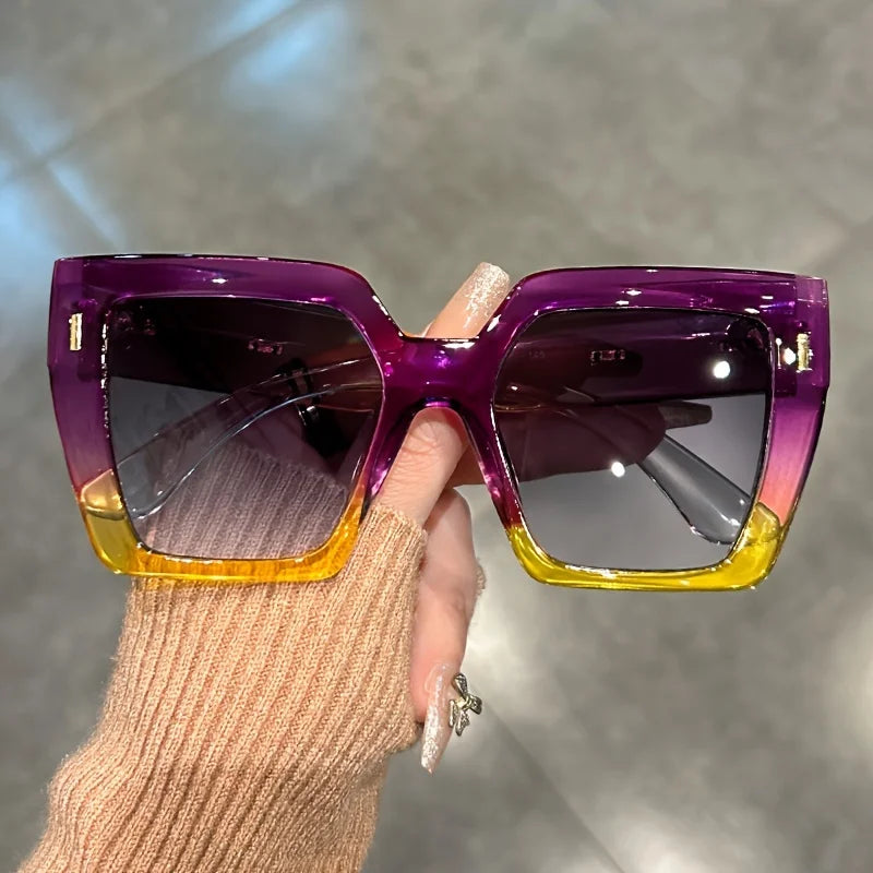 Purple gradient square sunglasses with gold accents being held by a hand.
