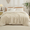 A beige soft muslin cotton blanket is draped over a white bed with white pillows and natural light.