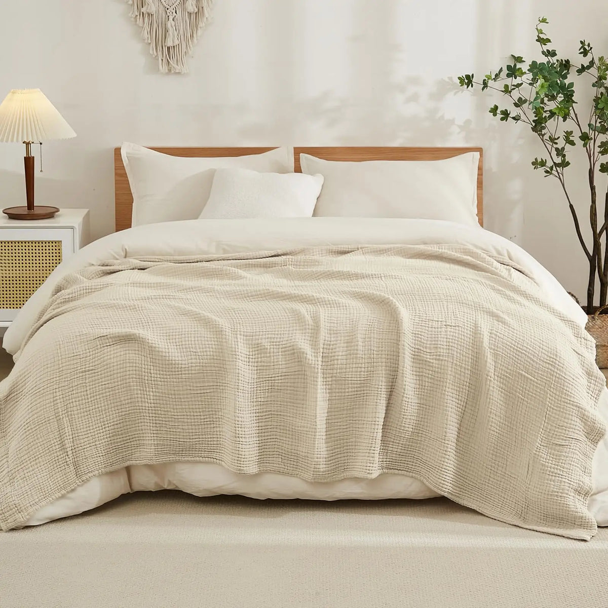 A beige soft muslin cotton blanket is draped over a white bed with white pillows and natural light.
