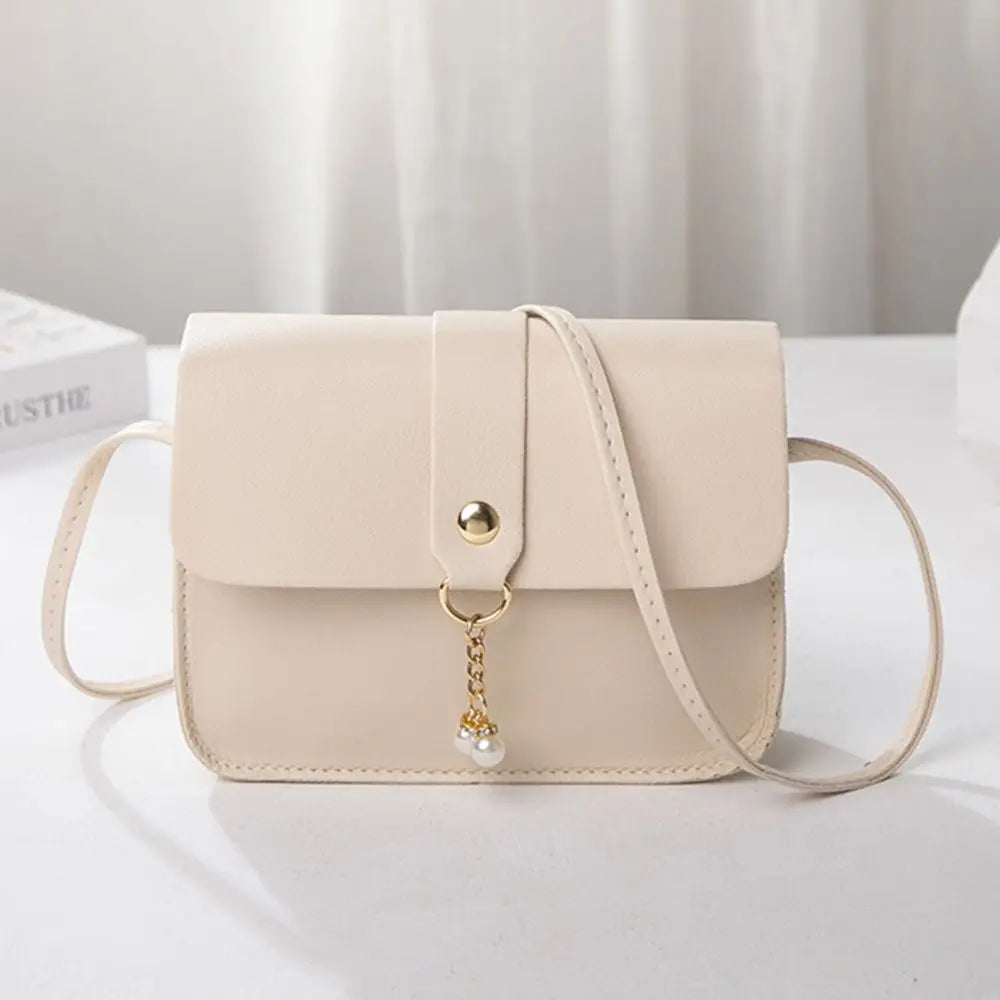 A beige compact crossbody handbag with a gold-tone chain charm and adjustable strap, shown on a white surface.