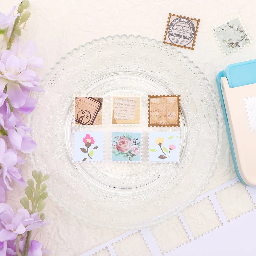 A clear glass plate with various square postage stamp paper punches and decorative stamps on a white background.