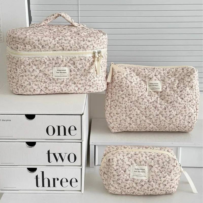 A set of three vintage floral quilted cosmetic bags in light pink and cream colors displayed on a white shelf.