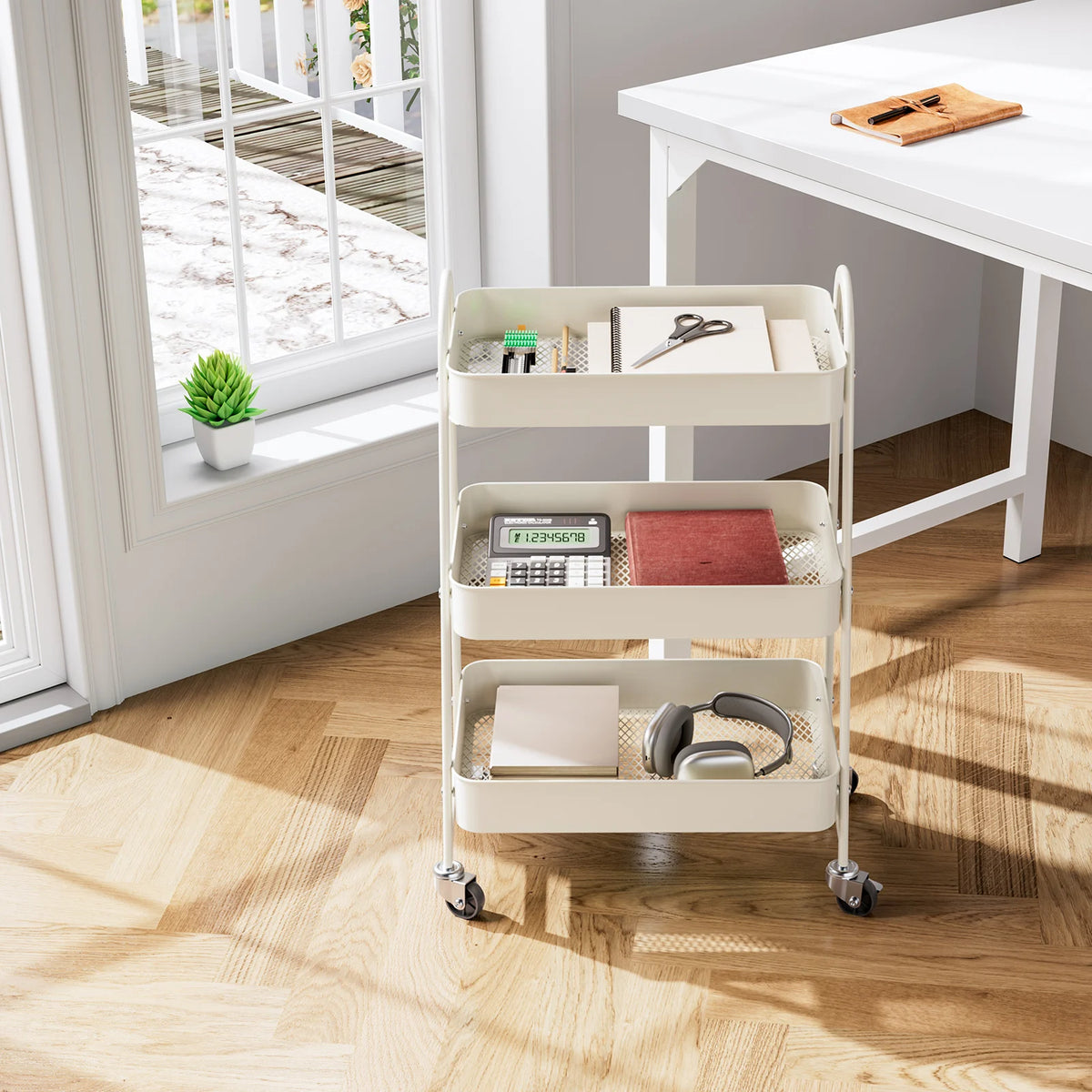 A white 3-tier rolling metal storage cart with lockable wheels holding various office supplies on a wooden floor.