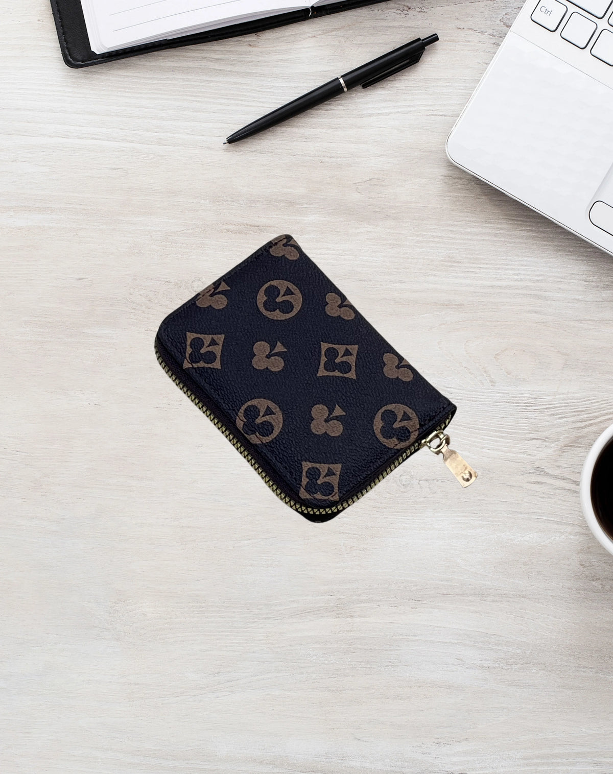 A dark patterned zippered card holder wallet resting on a light wooden surface next to a pen and a laptop.