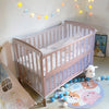 A light pink wooden baby crib with a white mattress and bedding is shown in a brightly lit room.