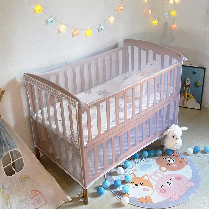 A light pink wooden baby crib with a white mattress and bedding is shown in a brightly lit room.