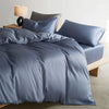 A smooth, slate blue luxury mulberry silk duvet cover set displayed on a bed with a neutral-toned lamp.