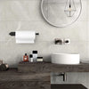 A stainless steel wall-mounted paper towel holder above a white vessel sink and mirror on a gray tiled wall.