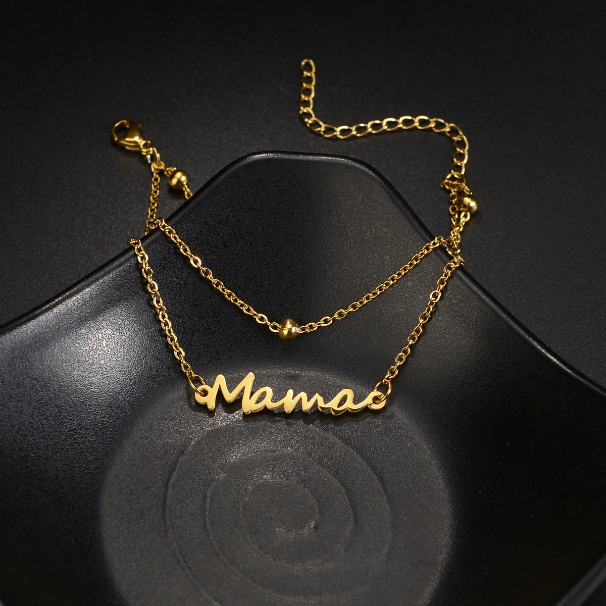 Gold double-layer name anklet spelling out Mama on a black surface