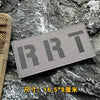 A rectangular gray tactical morale patch with the letters RRT embroidered on it, placed on a dark, rough stone surface.