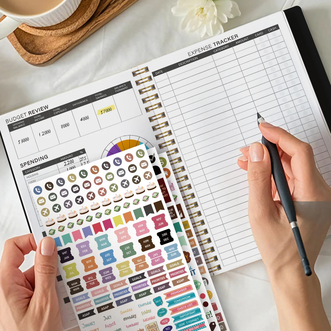 A hand is writing in an expense tracker budget planner with stickers and a pen on a white background.