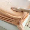 A hand is folding a peach-colored cotton fitted sheet with an elastic band on a white surface.