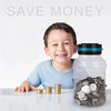 A smiling boy is sitting at a table with a clear glass jar filled with silver coins and a digital coin counting piggy bank.
