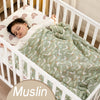 A baby is sleeping in a white crib wrapped in a green muslin swaddle blanket.