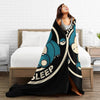 A woman is relaxing on a bed with a black and teal patterned Snorlax plush throw blanket.