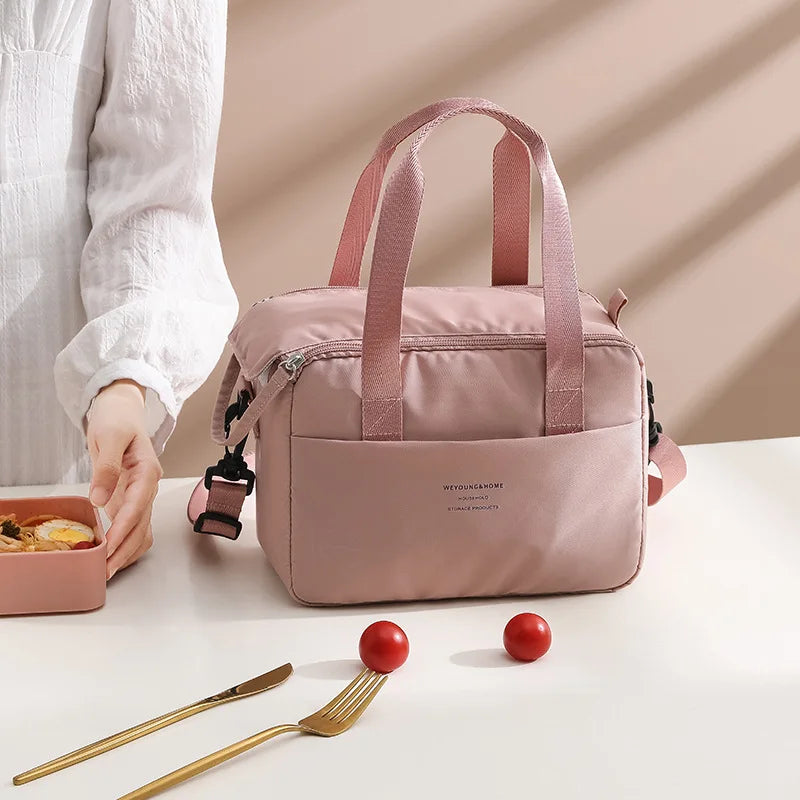 A pink compact thermal lunch tote bag with a canvas strap is shown on a white surface next to a fork.