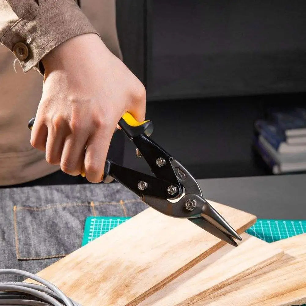 A hand is using heavy-duty aviation snips to cut a piece of wood on a workbench.