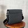 A dark gray patterned crossbody bag resting on a wooden stool against a light gray concrete background.