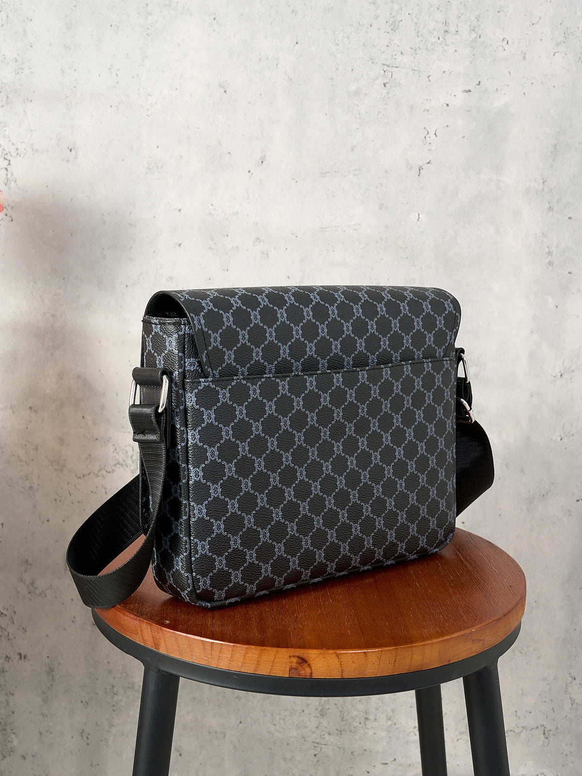 A dark gray patterned crossbody bag resting on a wooden stool against a light gray concrete background.