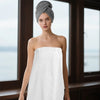 A woman wearing a gray turban stands in front of a window, holding a white microfiber bath wrap and hair towel set.
