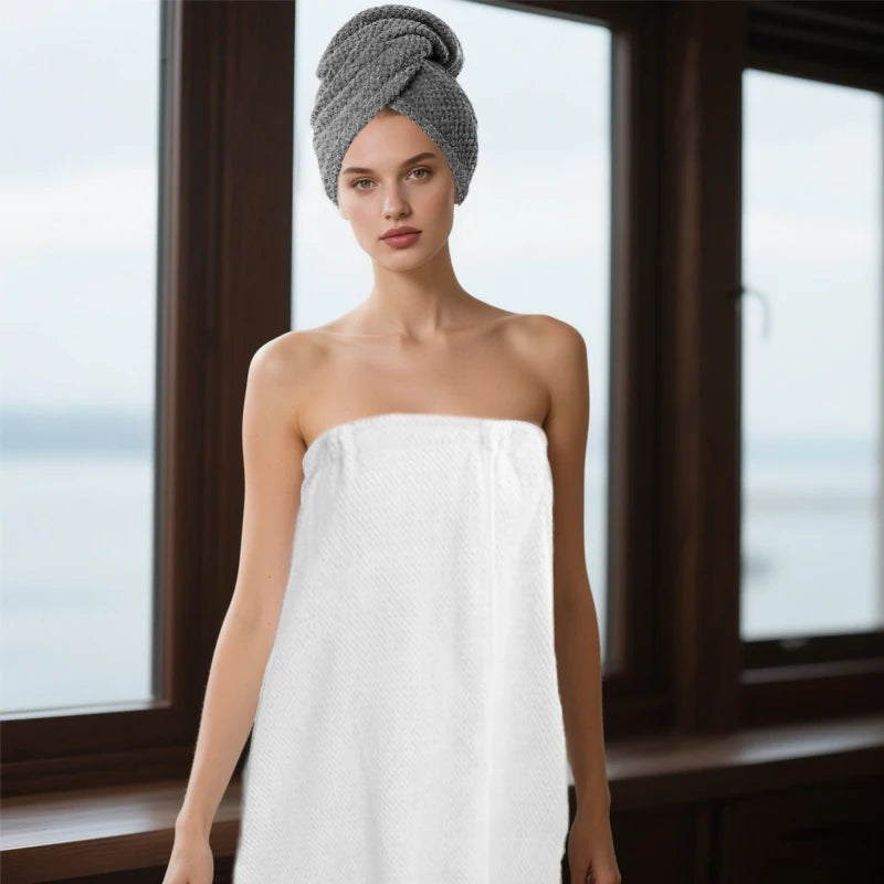 A woman wearing a gray turban stands in front of a window, holding a white microfiber bath wrap and hair towel set.