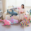 Eine Frau sitzt auf einem Sofa umgeben von großen Plüsch-Hai-Spielzeugen in rosa, blau und grau.