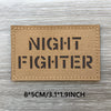 A rectangular tan fabric patch with the text NIGHT FIGHTER printed in black block letters on a weathered wooden surface.