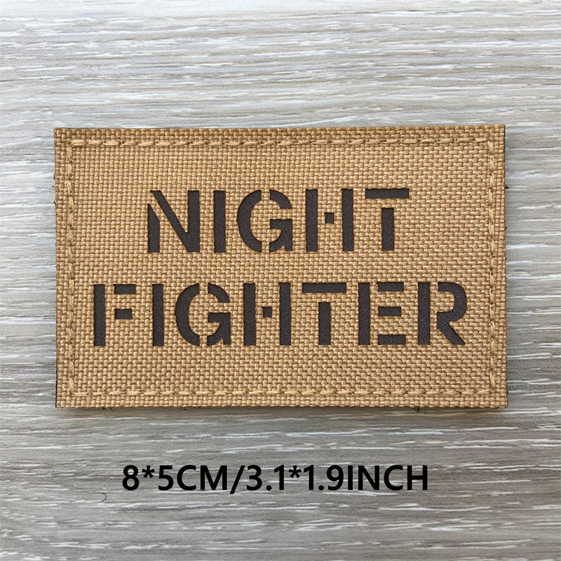A rectangular tan fabric patch with the text NIGHT FIGHTER printed in black block letters on a weathered wooden surface.