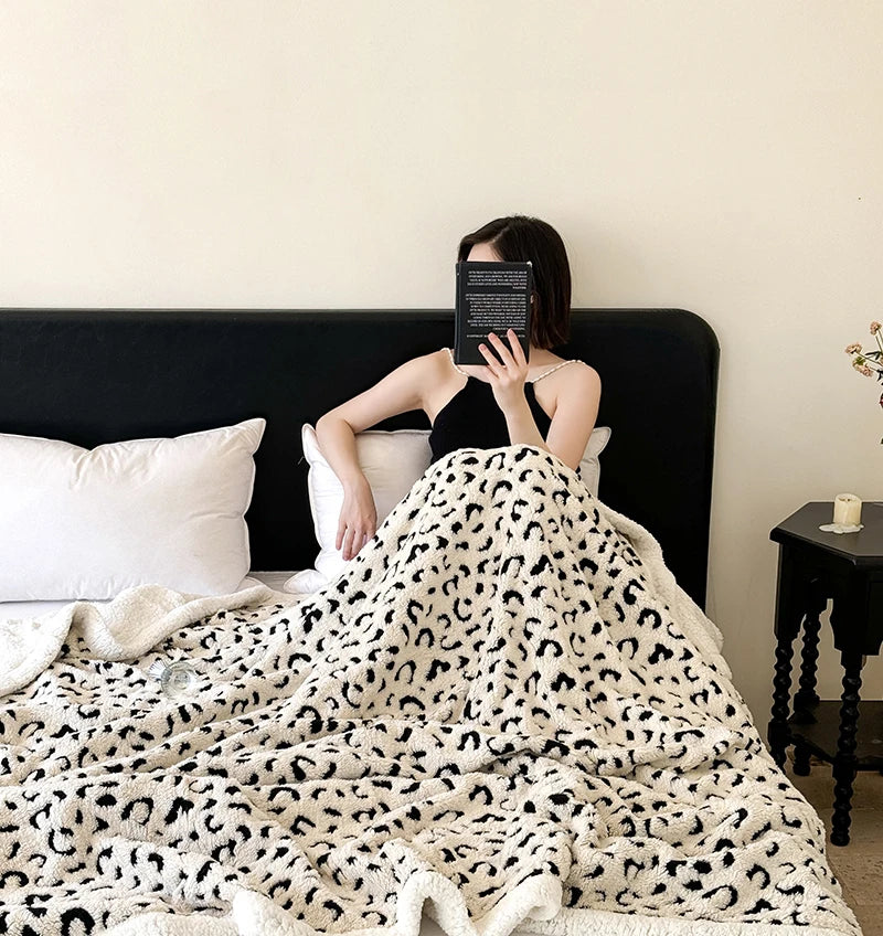 A woman is wrapped in a white and black leopard print plush jacquard throw blanket on a dark bed.