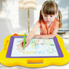 A young girl is drawing on a large yellow magnetic board with green markers.
