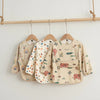 A beige waterproof long-sleeve art smock for toddlers with various nature and animal prints hanging on a wooden rack.